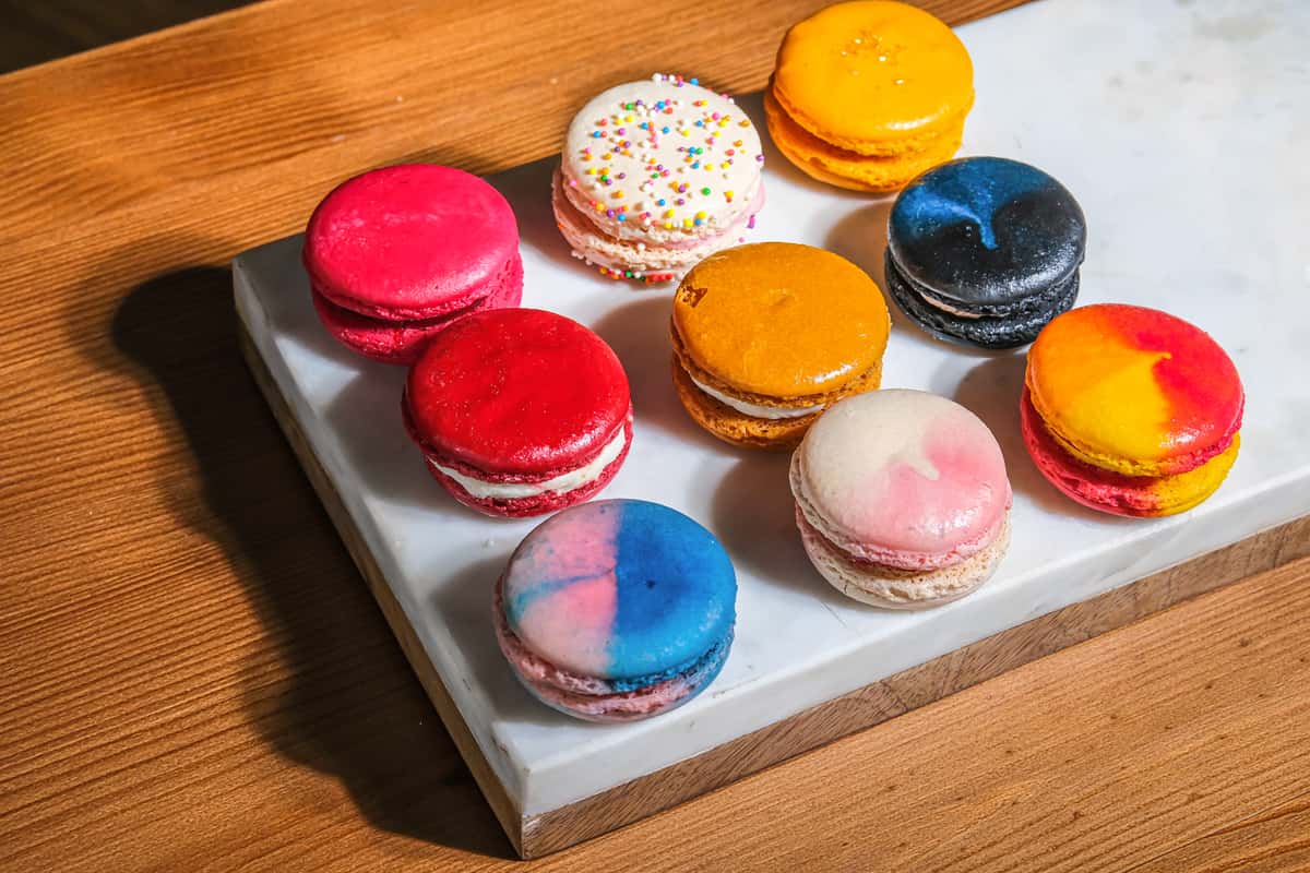 Colorful assorted macarons arranged on a white marble board, featuring vibrant pink, red, yellow, blue, and marbled shells on a wooden table