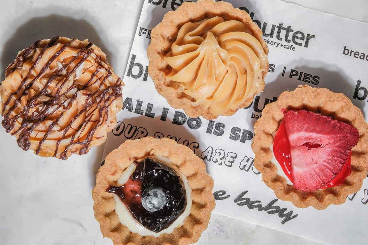 Assorted mini tarts topped with caramel, chocolate drizzle, blueberry, and strawberry on bakery-branded parchment paper.