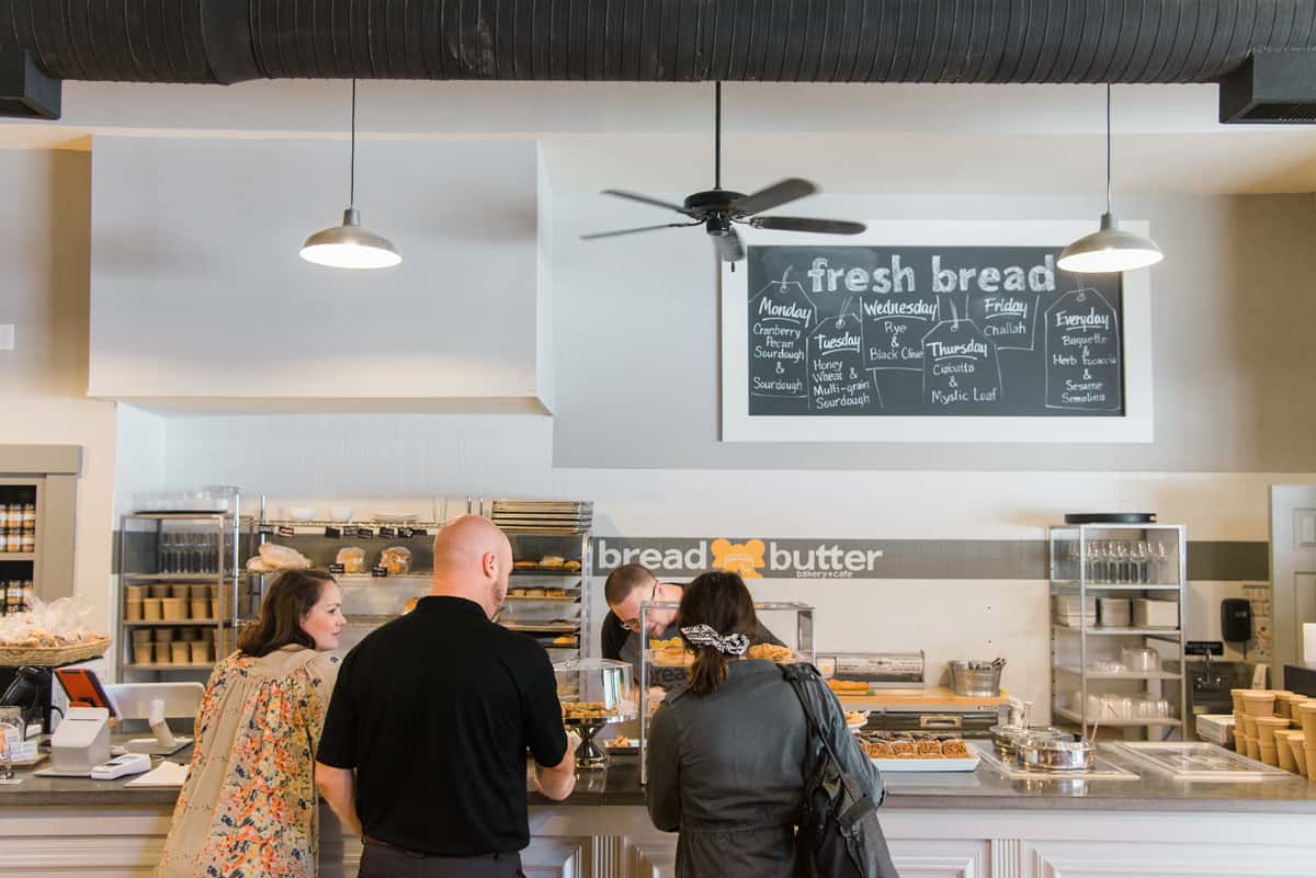 Customers ordering fresh baked goods at the Bread & Butter Bakery and Cafe counter beneath a chalkboard bread menu in a bright, welcoming bakery.