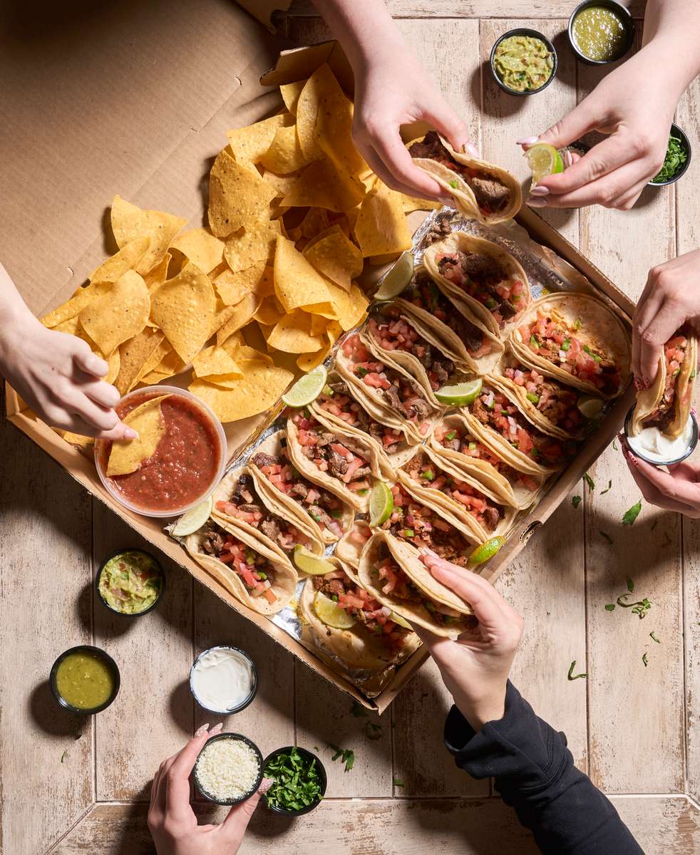 Overhead view of loaded gourmet hot dogs topped with fresh ingredients, served with kettle chips, dipping sauces, and citrus slices on a rustic table with guests sharing the meal.
