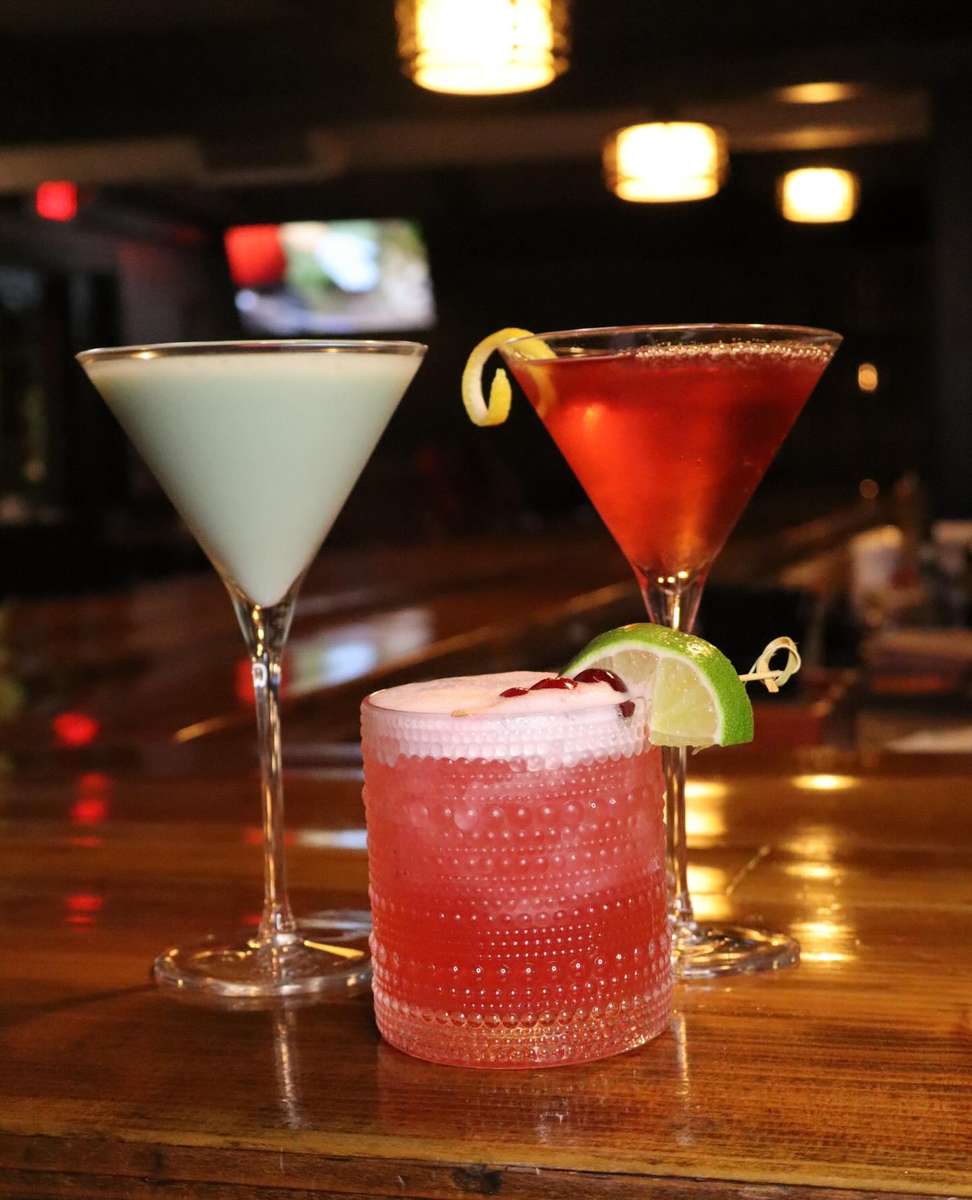 Three handcrafted cocktails on a polished bar—creamy martini, red martini with lemon twist, and frothy pink drink with lime garnish in a cozy pub setting.