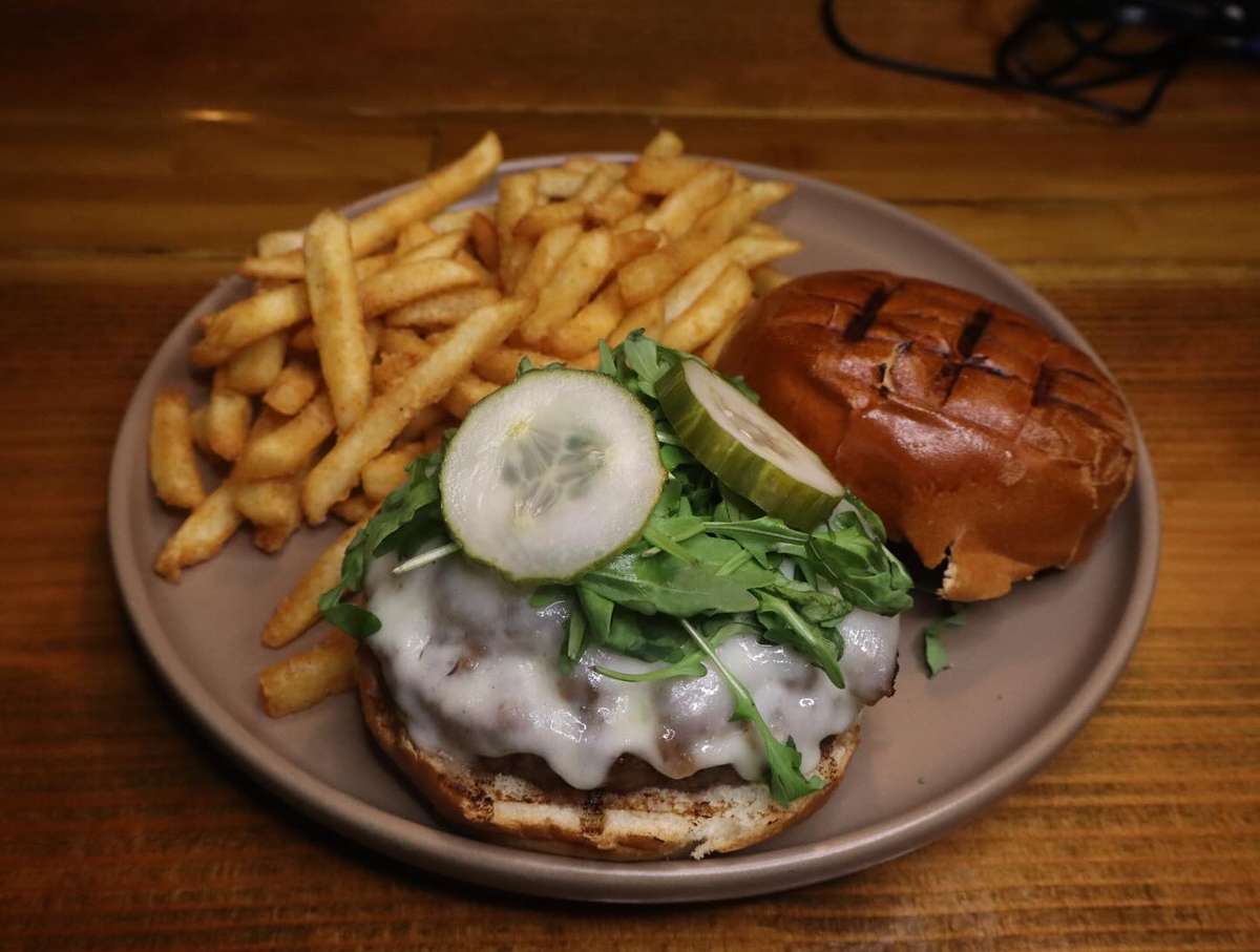 Cheeseburger with melted Swiss, arugula, and pickle on a toasted brioche bun, served with crispy French fries on a wooden bar.