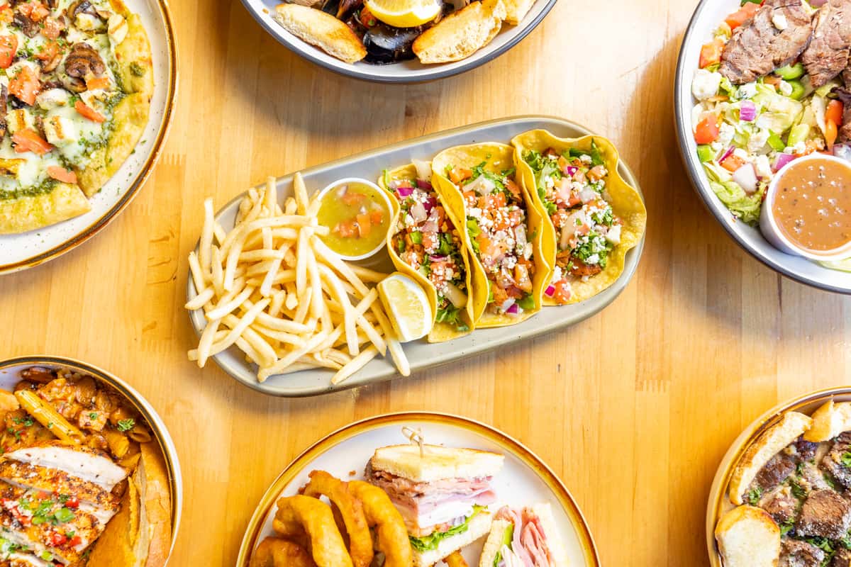 Vibrant restaurant platter featuring grilled chicken street tacos topped with cotija cheese, diced onions, cilantro, and salsa, served with a side of golden French fries and lemon wedges, surrounded by assorted dishes including flatbread pizza, mixed greens salad, seafood pasta, and deli-style sandwiches on a wooden table.
