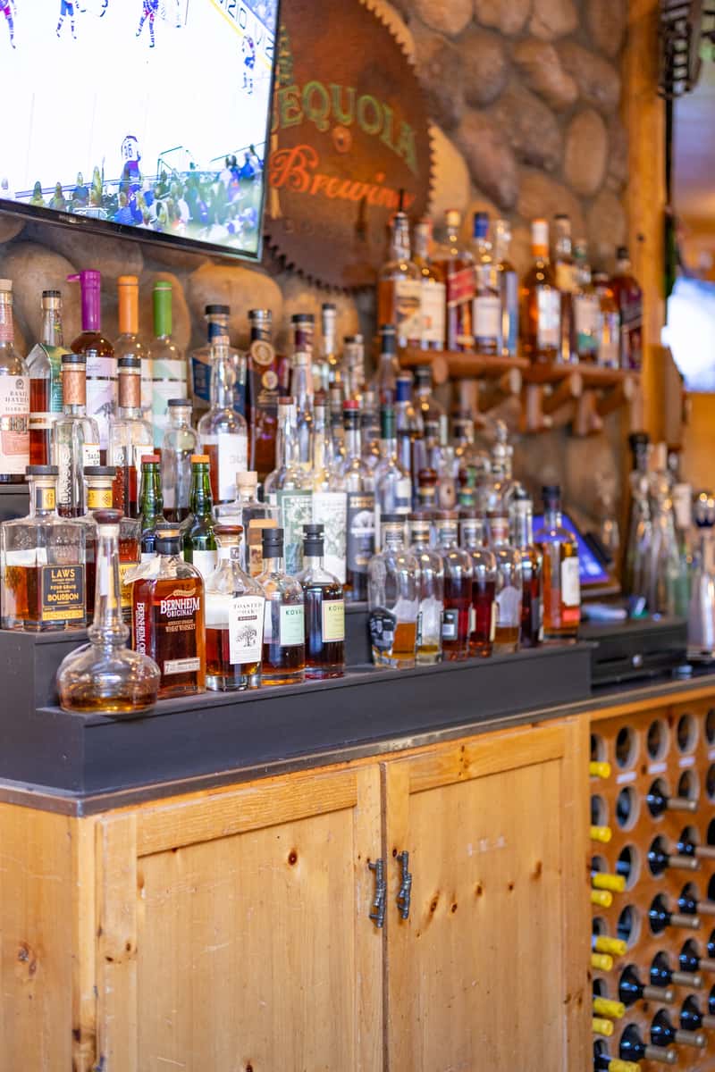 Expansive whiskey bar with a wide selection of premium spirits and bourbons neatly displayed on wooden shelving, set against a stone wall with Sequoia Brewing signage.