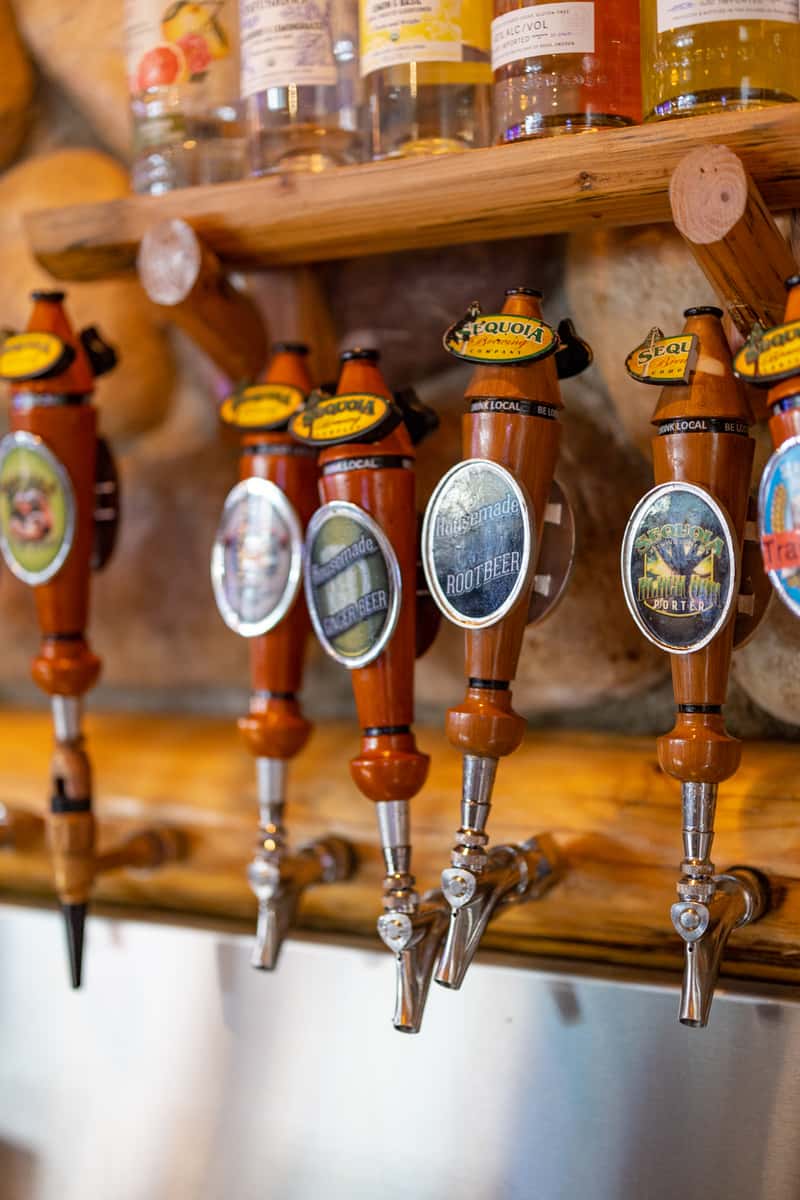 Close-up of Sequoia Brewing Company's custom beer tap handles, featuring housemade root beer and handcrafted porter, set against a rustic log and stone bar backdrop with liquor bottles above.
