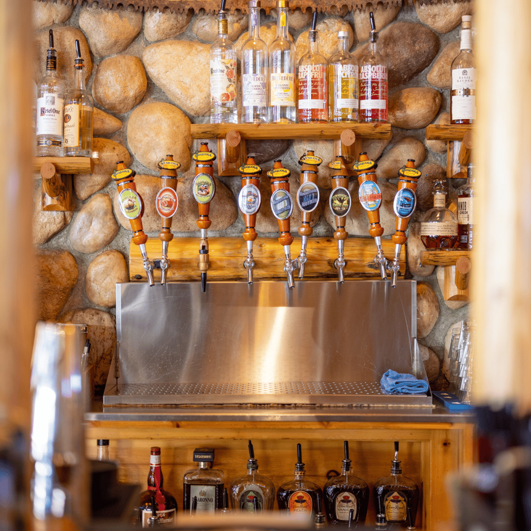 Rustic bar setup featuring a polished row of craft beer taps with custom wooden handles, set against a stone wall backdrop and surrounded by top-shelf liquor bottles and whiskey selections on wooden shelves.