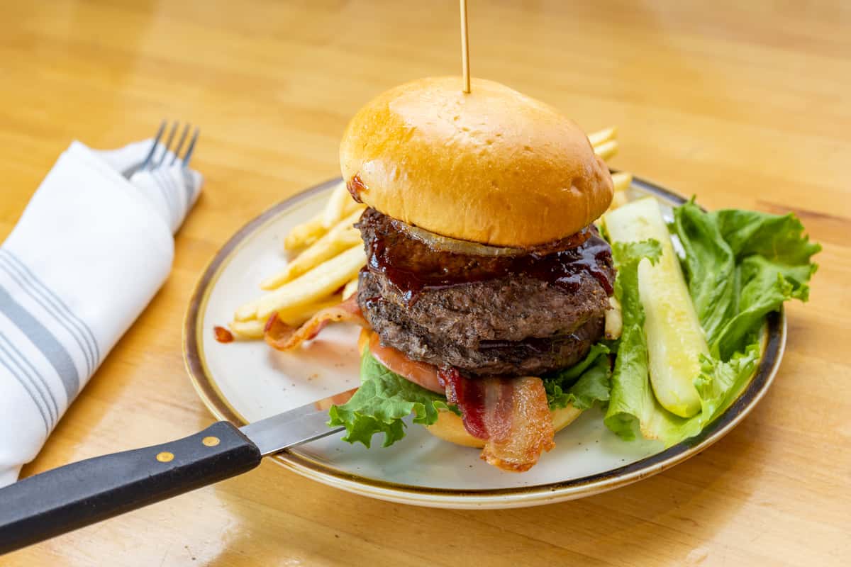 Hearty bacon cheeseburger stacked with lettuce, tomato, and crispy bacon on a brioche bun, served with golden fries and dill pickle spears on a white plate.