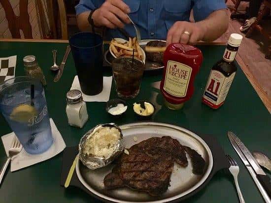 view of a plate with a steak and a baked potato. next to the plate is a glass of water, silverware, ketchup and a1 sauce