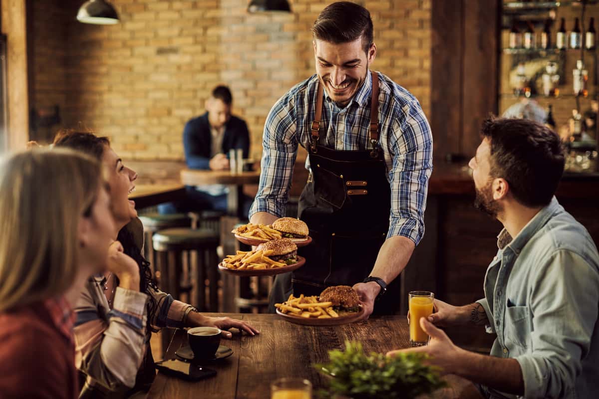 A smiling waiter serves plates of burgers and fries to a group of friends at a wooden table