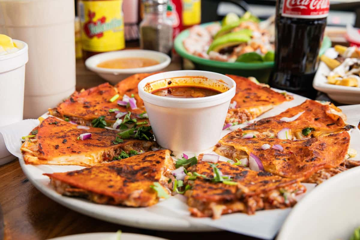 Vibrant red birria quesadillas with dipping consommé, topped with cilantro and onions, served on a table with other Mexican food at Pinches Tacos.