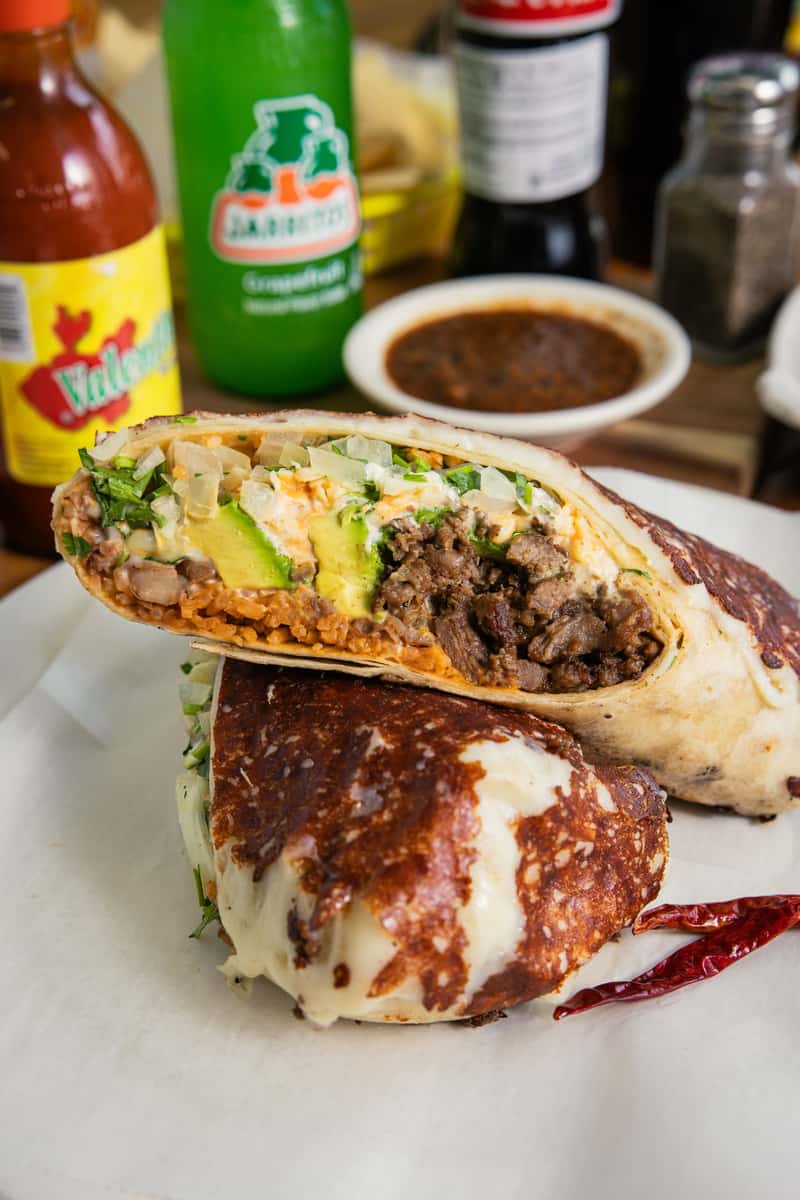 Pinches Tacos' hearty burrito, sliced open to show vibrant meat, avocado, and veggie fillings, featuring a melted, browned cheese exterior, with red hot sauce and a green Jarritos bottle in the background.