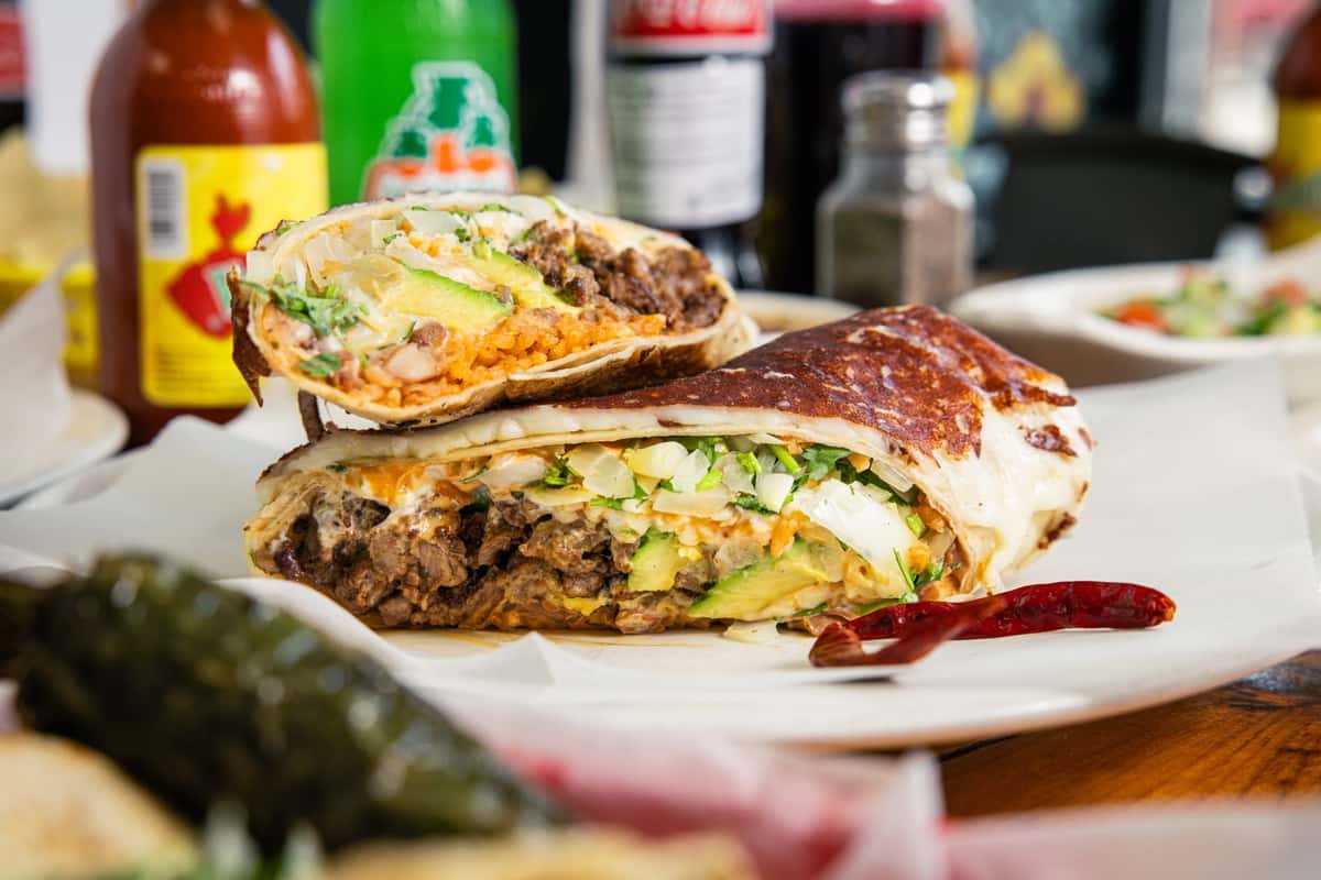 A vibrant Pinches Tacos burrito, cut open to reveal carne asada, green avocado, orange rice, and fresh vegetables, is plated with red dried chilies and a hot sauce bottle in the background.