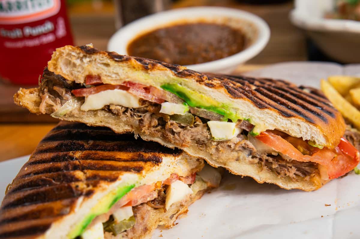 Pinches Tacos' vibrant grilled torta, showcasing shredded meat, green avocado, red tomato, and white cheese, with a red Jarritos drink.