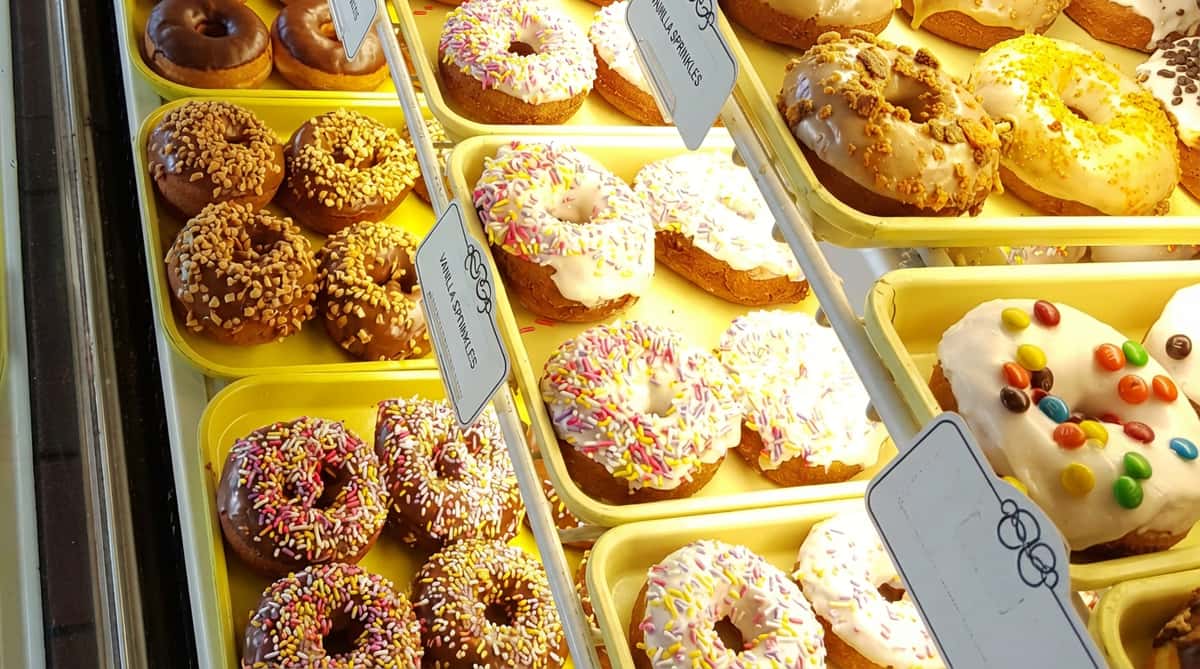 Assorted donuts in a display case; some with chocolate and nuts, vanilla with multi-colored sprinkles, and white icing with M&M's.