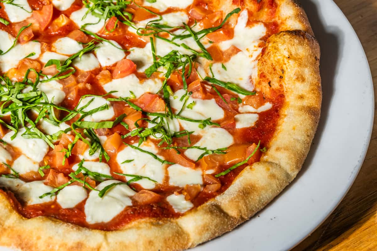 Close-up of a wood-fired Margherita pizza topped with fresh mozzarella, diced tomatoes, basil strips, and rich tomato sauce on a golden, blistered crust. A vibrant and classic Italian-American favorite.