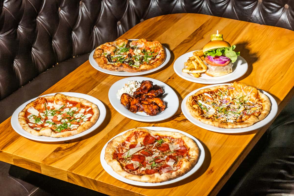Variety of wood-fired pizzas, saucy barbecue wings with coleslaw, and a classic cheeseburger with fries arranged on a rustic wooden table in a tufted leather booth setting. A hearty American grill spread featuring bold flavors and casual dining favorites.