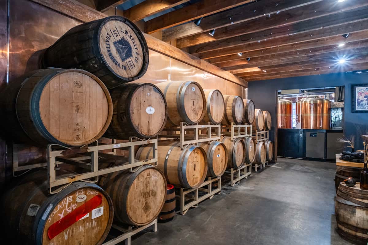 Rustic brewery barrel room featuring stacked wooden aging barrels and a view into the brewing area with polished copper tanks. Warm lighting and exposed wooden beams enhance the artisanal, craft brewery ambiance.