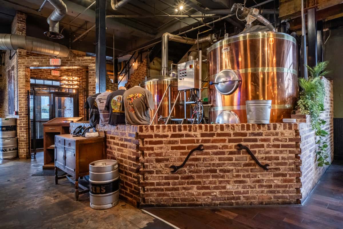 Interior of a craft brewery with large polished copper brewing tanks set behind a red brick partition. The industrial-style space features exposed ductwork, rustic wooden floors, and a merchandise area displaying branded t-shirts. A stylish fusion of vintage charm and modern brewery aesthetics.