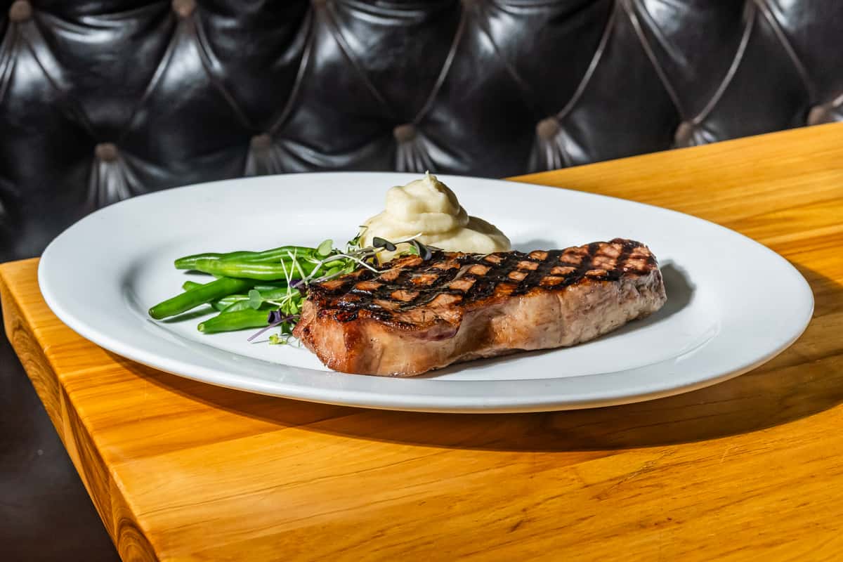 Juicy grilled New York strip steak with perfect char lines, served with buttery mashed potatoes and crisp sautéed green beans, garnished with fresh microgreens. A refined steakhouse dish presented on a white plate against a rustic wood table and tufted leather booth backdrop.