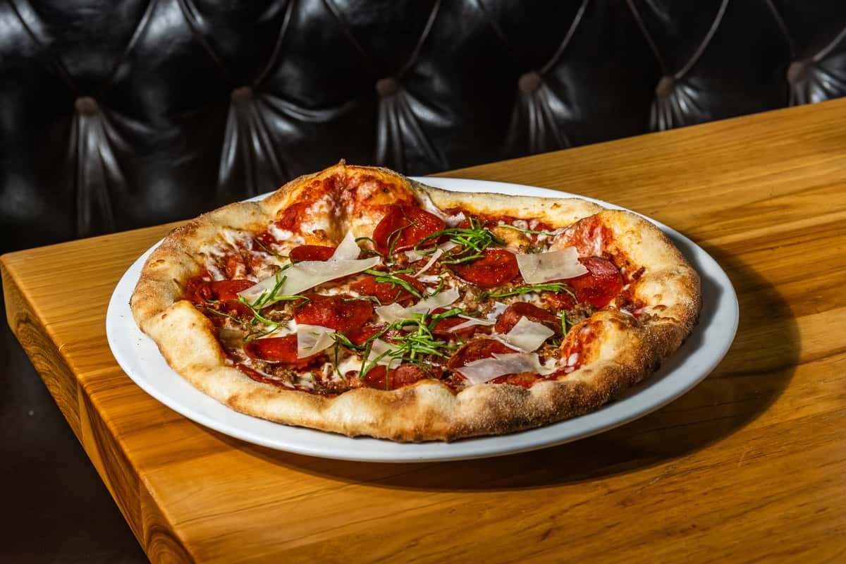 Wood-fired pepperoni pizza topped with shaved parmesan, tomato sauce, melted mozzarella, and fresh basil, served on a crisp, golden crust. A flavorful classic perfect for Italian-American cuisine lovers.