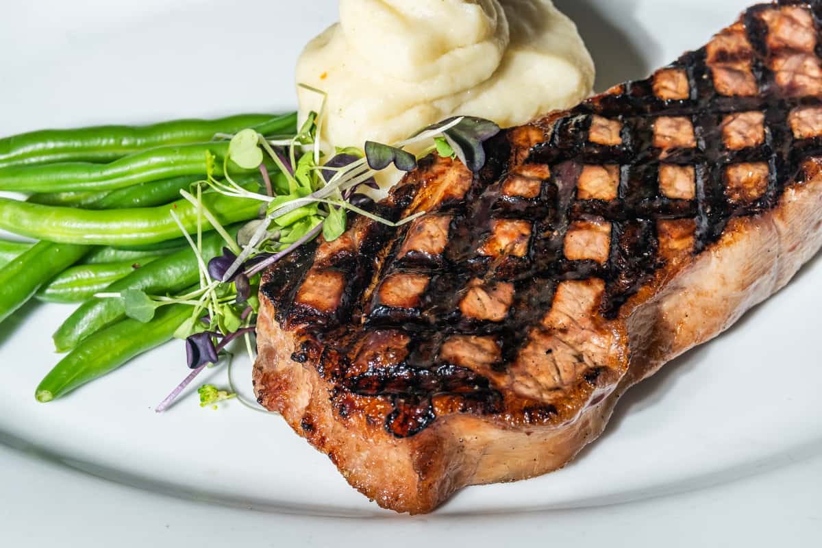 Chargrilled New York strip steak with crosshatch grill marks, served with garlic mashed potatoes, tender green beans, and fresh microgreens for garnish. A hearty steakhouse favorite with rustic elegance.