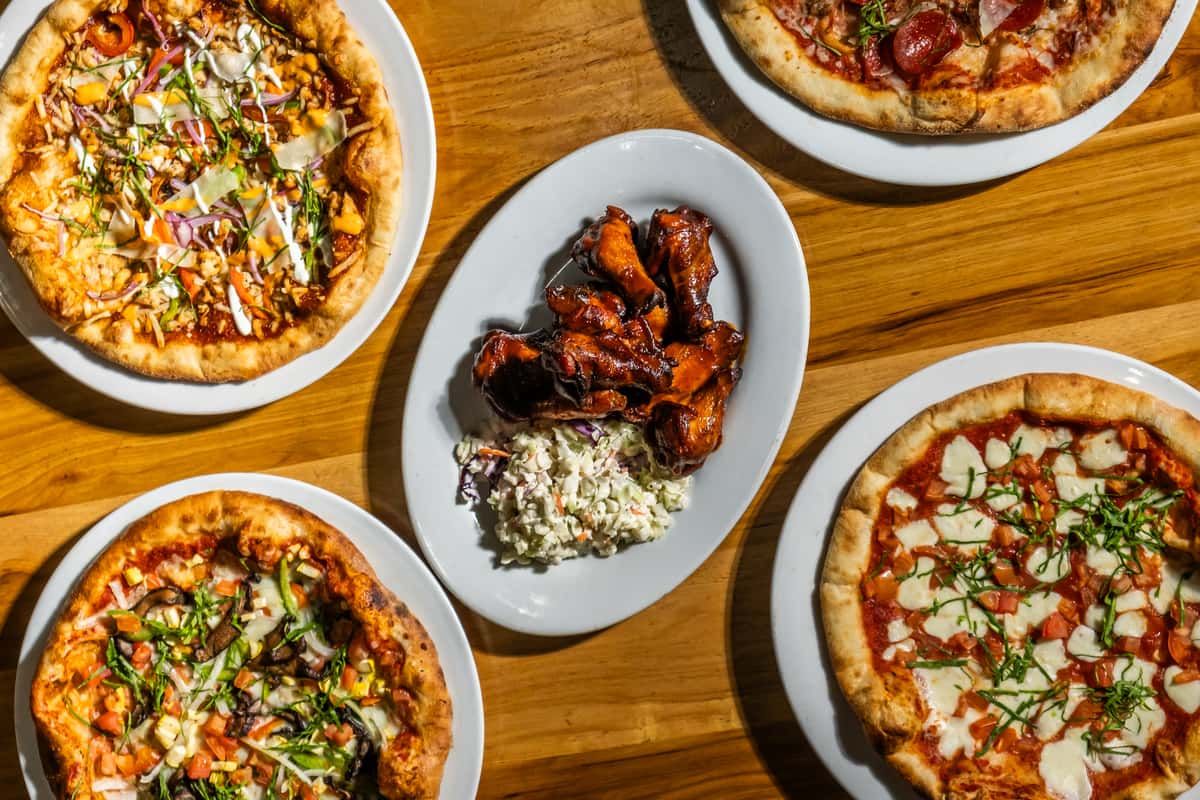 Top-down view of assorted wood-fired pizzas with toppings like pepperoni, mozzarella, fresh basil, and vegetables, centered around a plate of saucy barbecue wings and creamy coleslaw. A colorful and crave-worthy selection of American-Italian pub fare on a rustic wooden table.