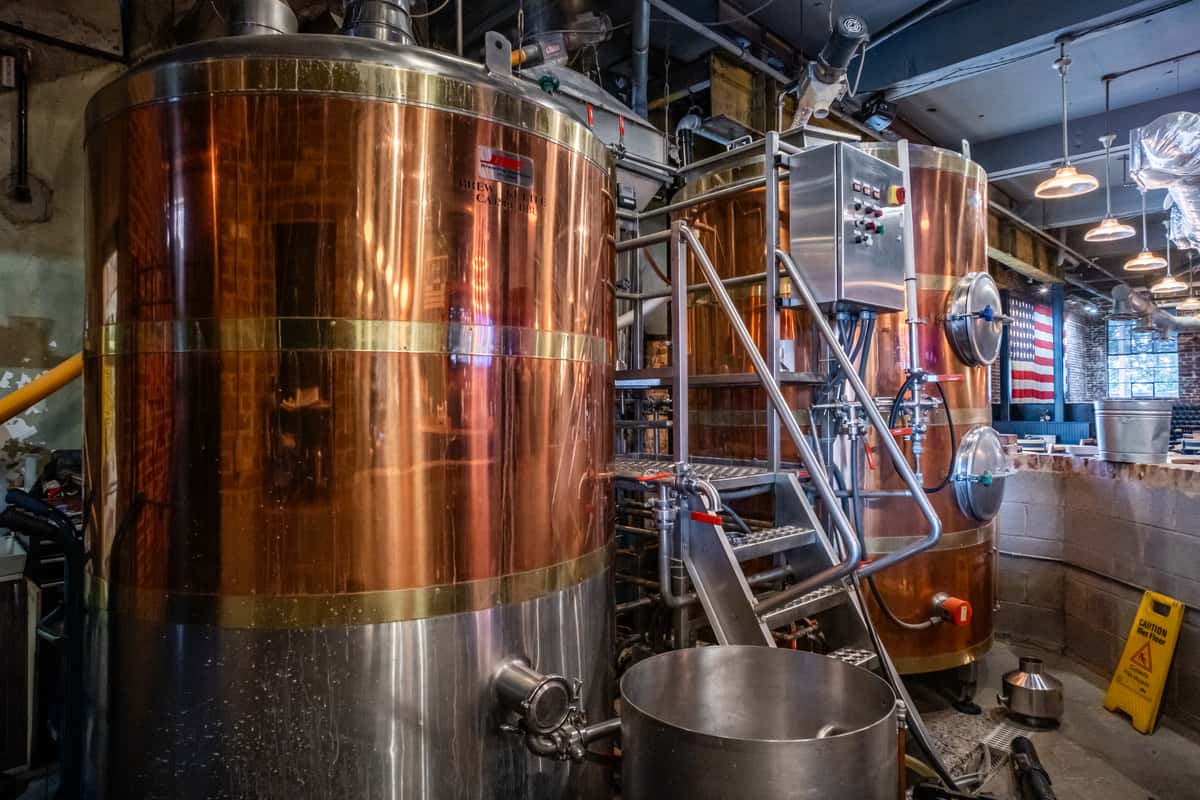 Close-up view of large copper brewing tanks and stainless steel equipment inside Max Lager's brewery. The industrial setup, surrounded by exposed pipes and pendant lighting, highlights the craft beer brewing process in a rustic, American brewpub setting.