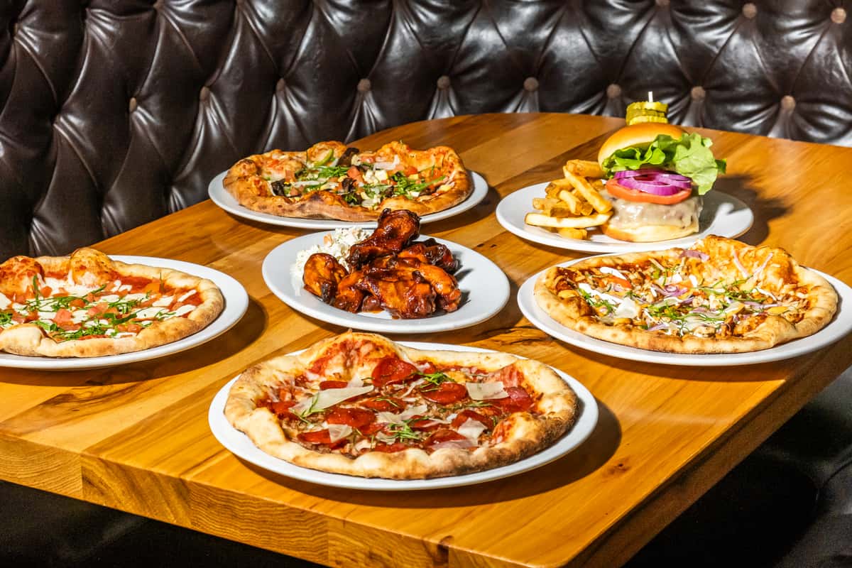 Assortment of handcrafted pizzas, saucy chicken wings, and a stacked cheeseburger with fries on a rustic wooden table in a cozy leather booth setting. The spread includes pepperoni pizza with parmesan, veggie pizza with mushrooms and peppers, margherita pizza, and barbecue chicken pizza, creating a vibrant mix of Italian-American comfort food.