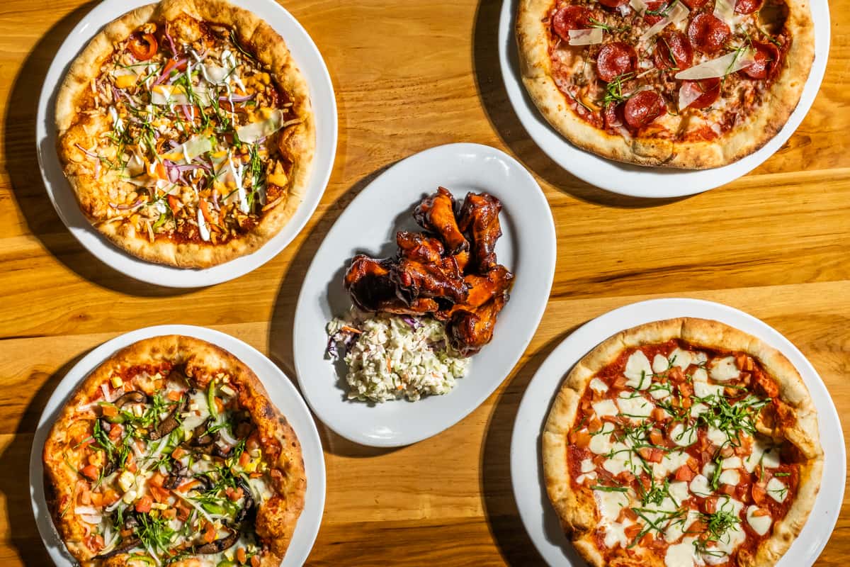 Overhead view of a wooden table featuring four artisanal pizzas with various toppings, including pepperoni, fresh mozzarella, basil, mushrooms, onions, and shaved parmesan. At the center is a platter of glazed chicken wings paired with creamy coleslaw, highlighting a casual dining experience with a rustic Italian-American flair.