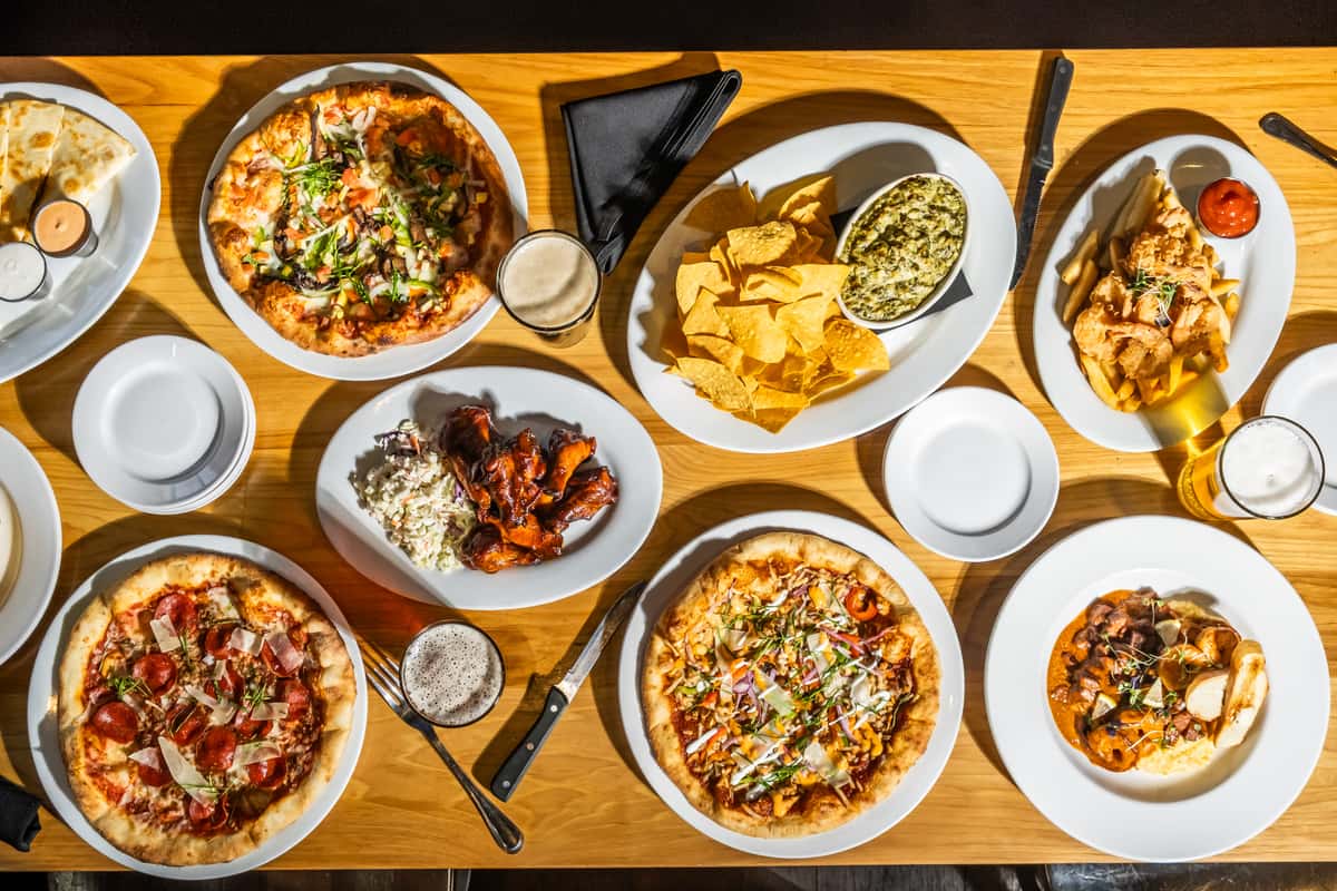 Overhead spread of wood-fired pizzas, saucy wings with coleslaw, chicken quesadillas, spinach artichoke dip with tortilla chips, fried shrimp, and creamy pasta, paired with craft beers. A vibrant, shareable feast showcasing the variety and bold flavors of American grill and brewpub cuisine.