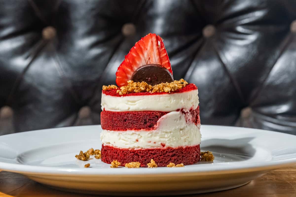 Elegant red velvet layer cake with cream cheese frosting, garnished with a sliced strawberry, chocolate coin, and crunchy crumble topping. A rich and visually striking dessert served on a white plate with a tufted leather booth backdrop.