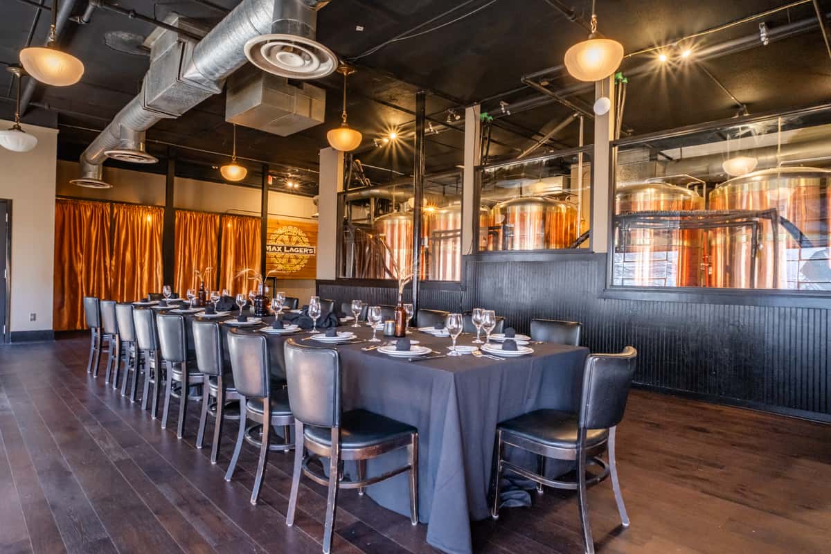 Elegant private dining room at Max Lager's featuring a long banquet-style table with black linens, formal place settings, and a view of copper brewing tanks through glass panels. A refined space blending industrial brewery elements with upscale event ambiance.