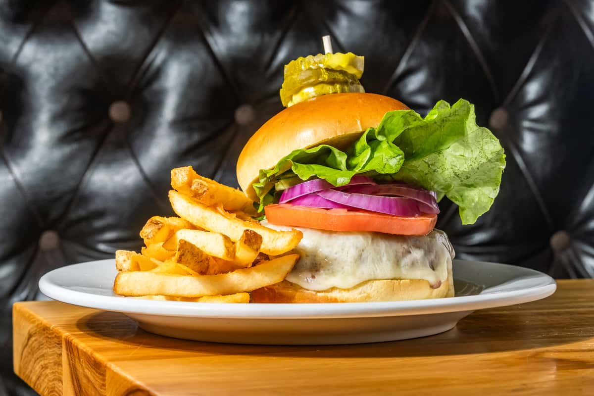 Juicy cheeseburger stacked with lettuce, tomato, red onion, and melted cheese on a toasted bun, topped with pickle slices and served with golden French fries. A classic American comfort food plate presented in a cozy, upscale setting.