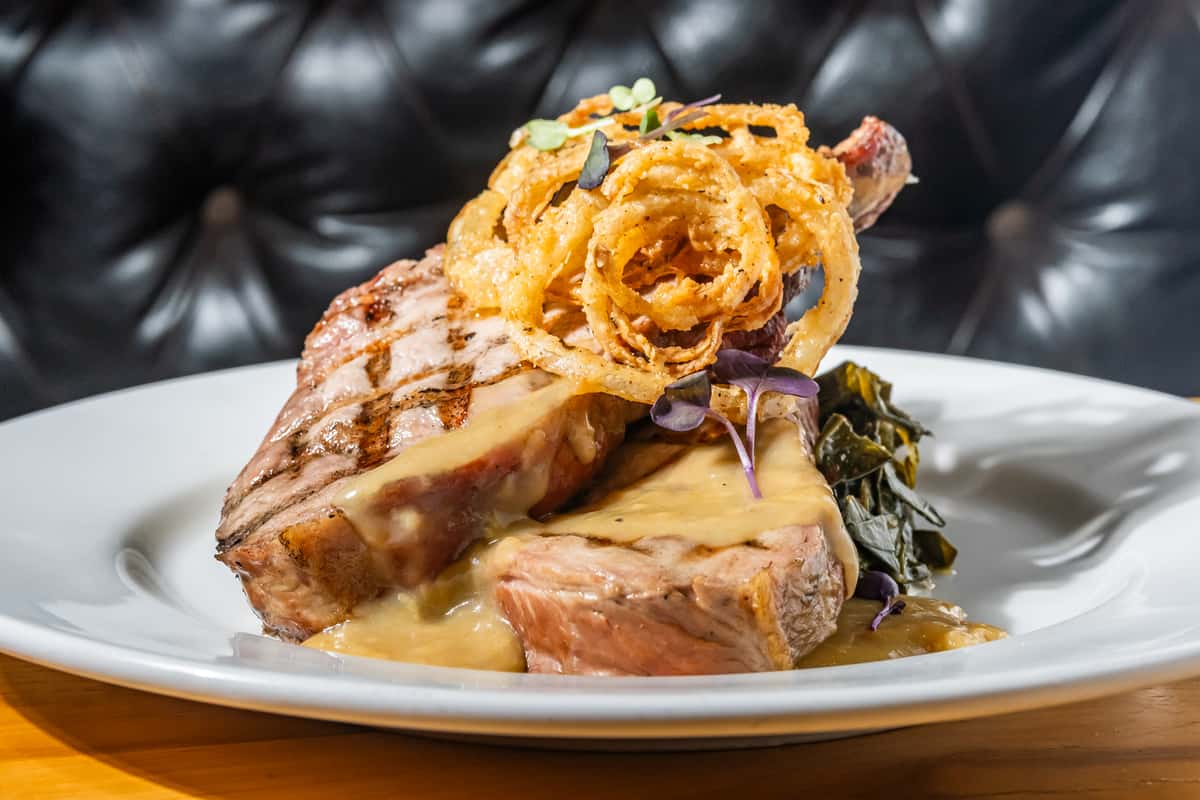 Bone-in grilled pork chop smothered in savory gravy, topped with crispy fried onions and microgreens, plated with braised greens. A Southern-inspired entrée with bold, comforting flavors.