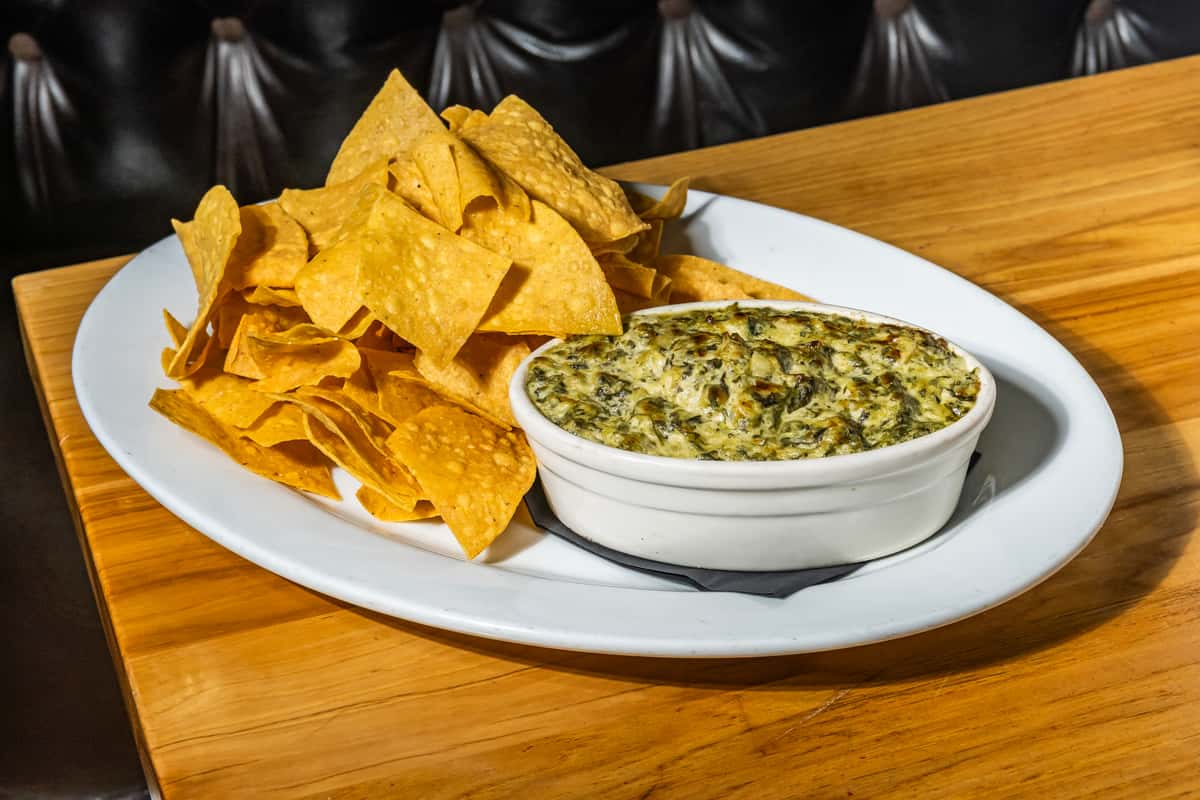 Warm spinach and artichoke dip served in a ceramic ramekin alongside a generous portion of crispy tortilla chips. A popular appetizer combining creamy, cheesy goodness with crunchy dippable chips, perfect for sharing at a casual American grill.