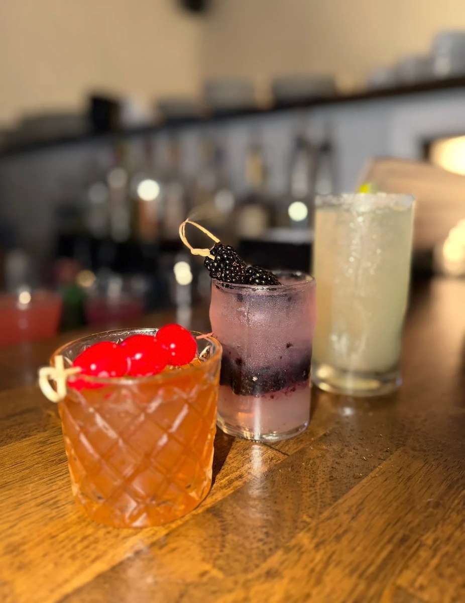 Three handcrafted cocktails on a wooden bar, including an Old Fashioned with cherries, a blackberry-infused vodka drink, and a classic margarita with a salted rim, perfect for an upscale lounge or Southern restaurant bar.