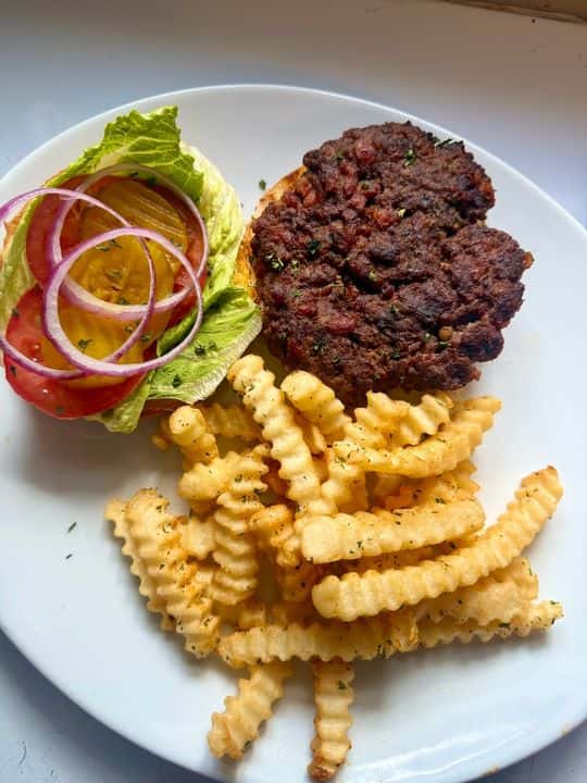 Classic hamburger steak patty with crinkle-cut fries, served with lettuce, tomato, pickles, and red onion on a white plate.