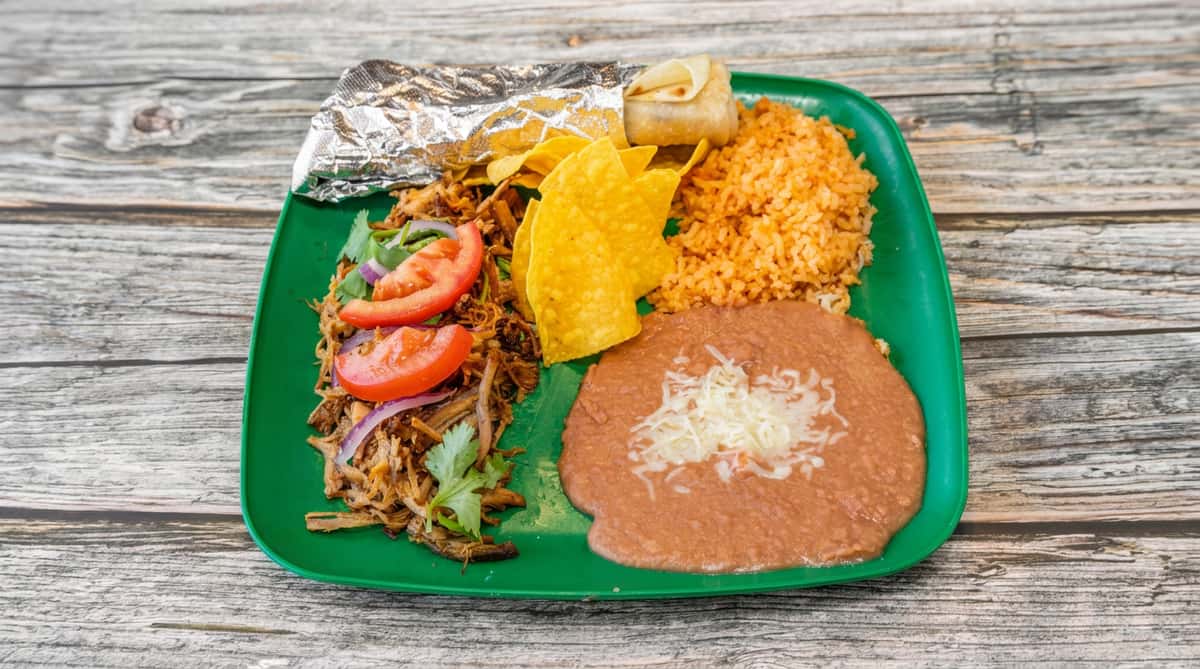 Mexican carnitas plate with shredded pork, tomato, red onion, cilantro, rice, refried beans with cheese, chips, and a mini burrito.
