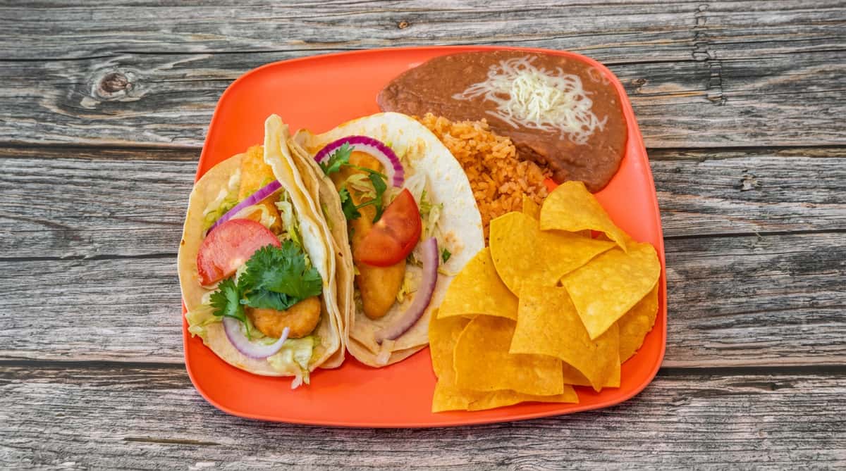 Fish tacos with shredded lettuce, red onion, tomato, and cilantro, served with refried beans, rice, and tortilla chips.