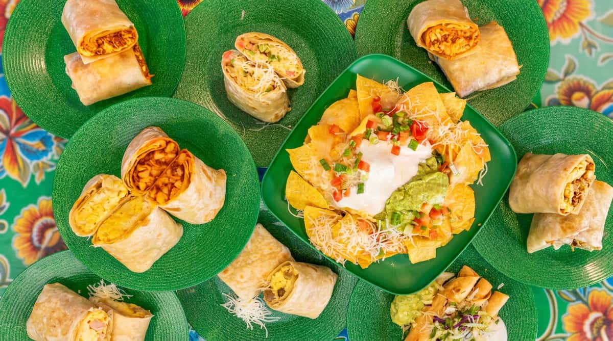 An assortment of burritos, nachos with guacamole and sour cream, and taquitos on green plates.