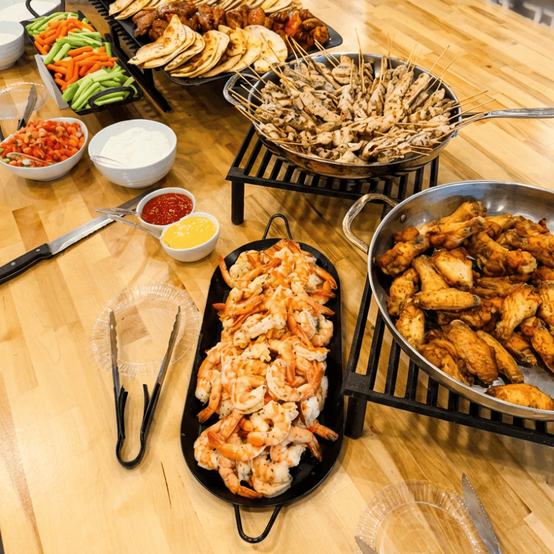 Catering buffet spread with shrimp platter, chicken wings, grilled skewers, tacos, fresh vegetables, and dipping sauces arranged on a wooden table for party catering.
