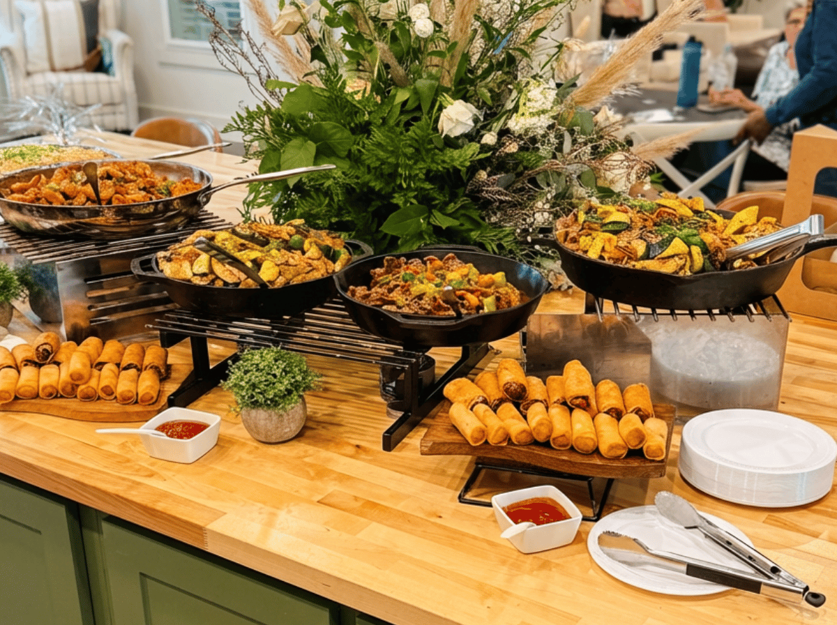 Catering buffet display with assorted hot dishes, fried appetizers, and crispy egg rolls served in cast-iron skillets on a wooden table with decorative greenery centerpiece.