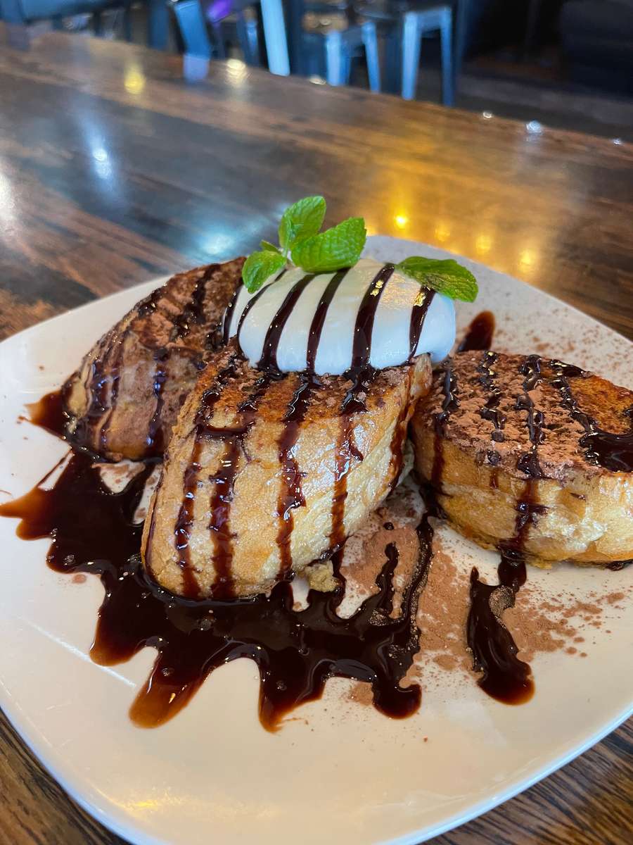 A decadent dessert featuring thick slices of golden-brown French toast drizzled with rich chocolate sauce and topped with a scoop of vanilla ice cream. Finished with whipped cream and a drizzle of chocolate, this indulgent treat offers the perfect balance of warm, sweet flavors and creamy textures.