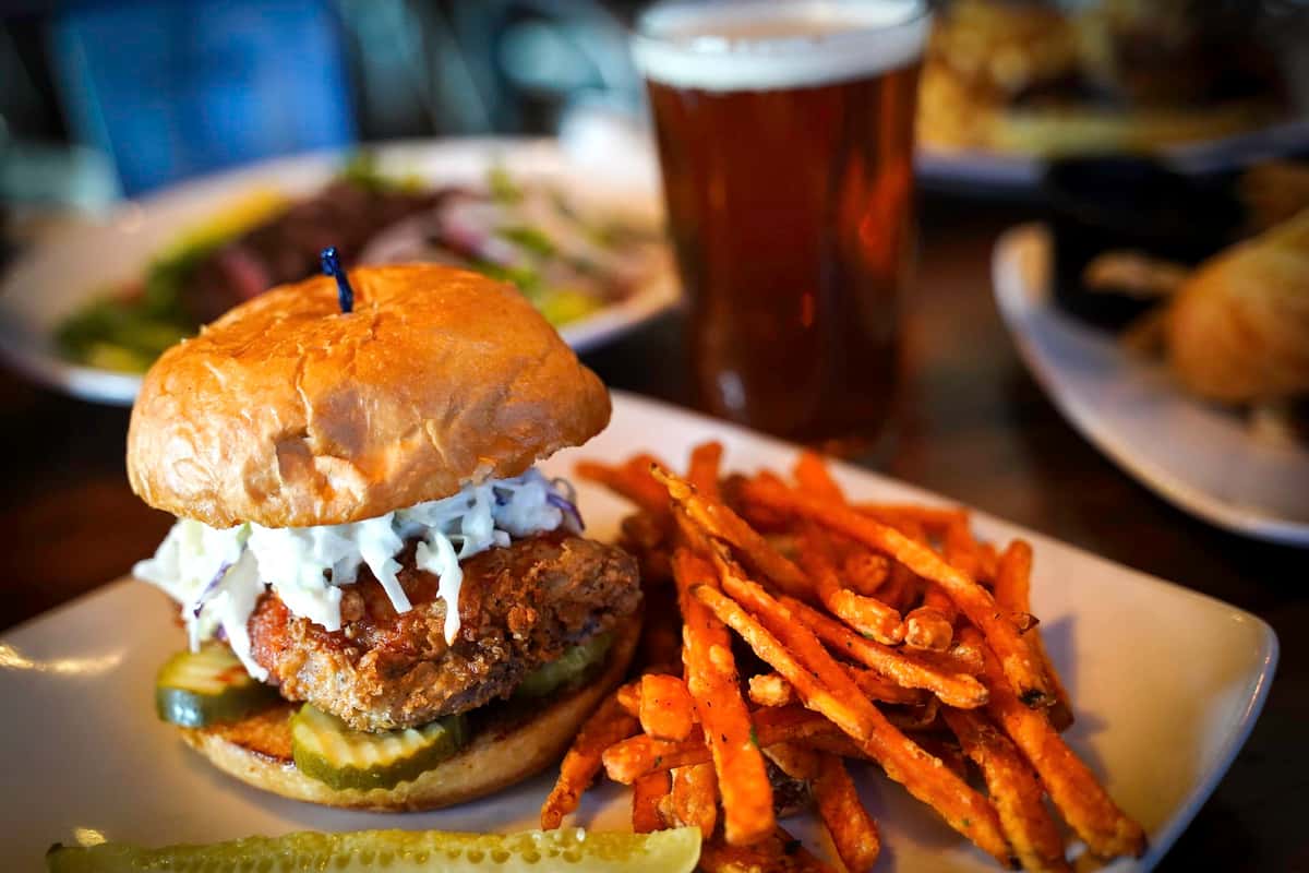 A juicy gourmet burger stacked with melted cheese, fresh toppings, and a creamy sauce sits on a toasted bun alongside a generous serving of crispy sweet potato fries. In the background, a cold pint of beer completes the scene, creating the perfect pairing for a hearty, satisfying meal at a casual grill or pub.