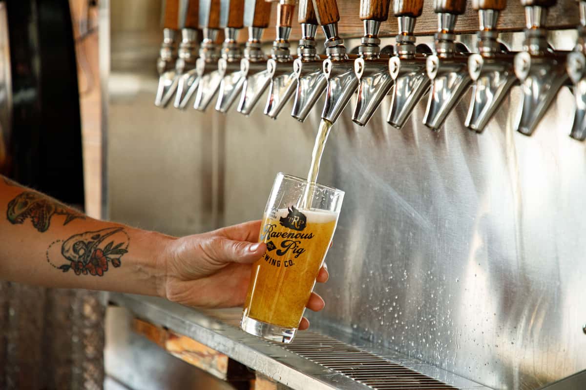 A bartender pours a freshly crafted golden beer from a row of stainless-steel taps into a branded pint glass. Foam forms at the top as the beer fills the glass, while the blurred tap handles and bar backdrop create a clean, modern brewery atmosphere.