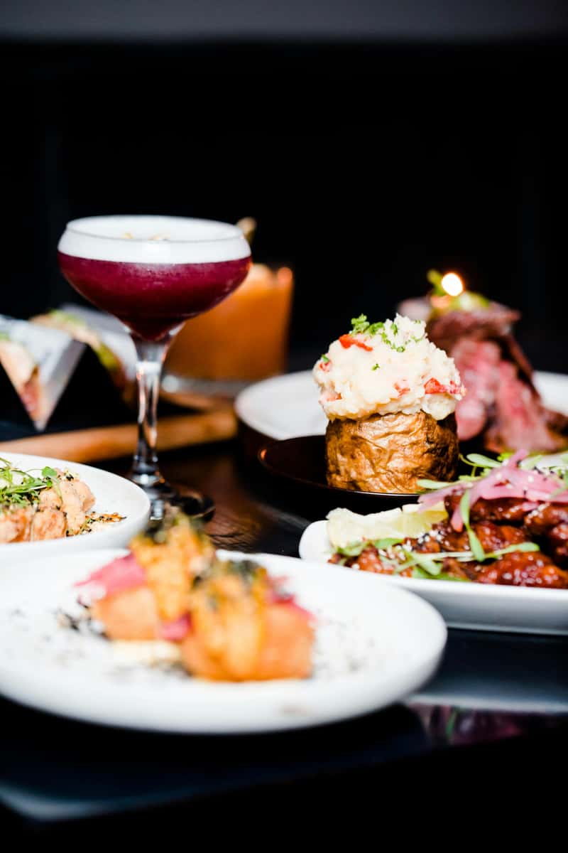 Elegant dinner spread featuring a berry cocktail, baked potato with crab, ribs, sushi, and gourmet small plates on a dark table.