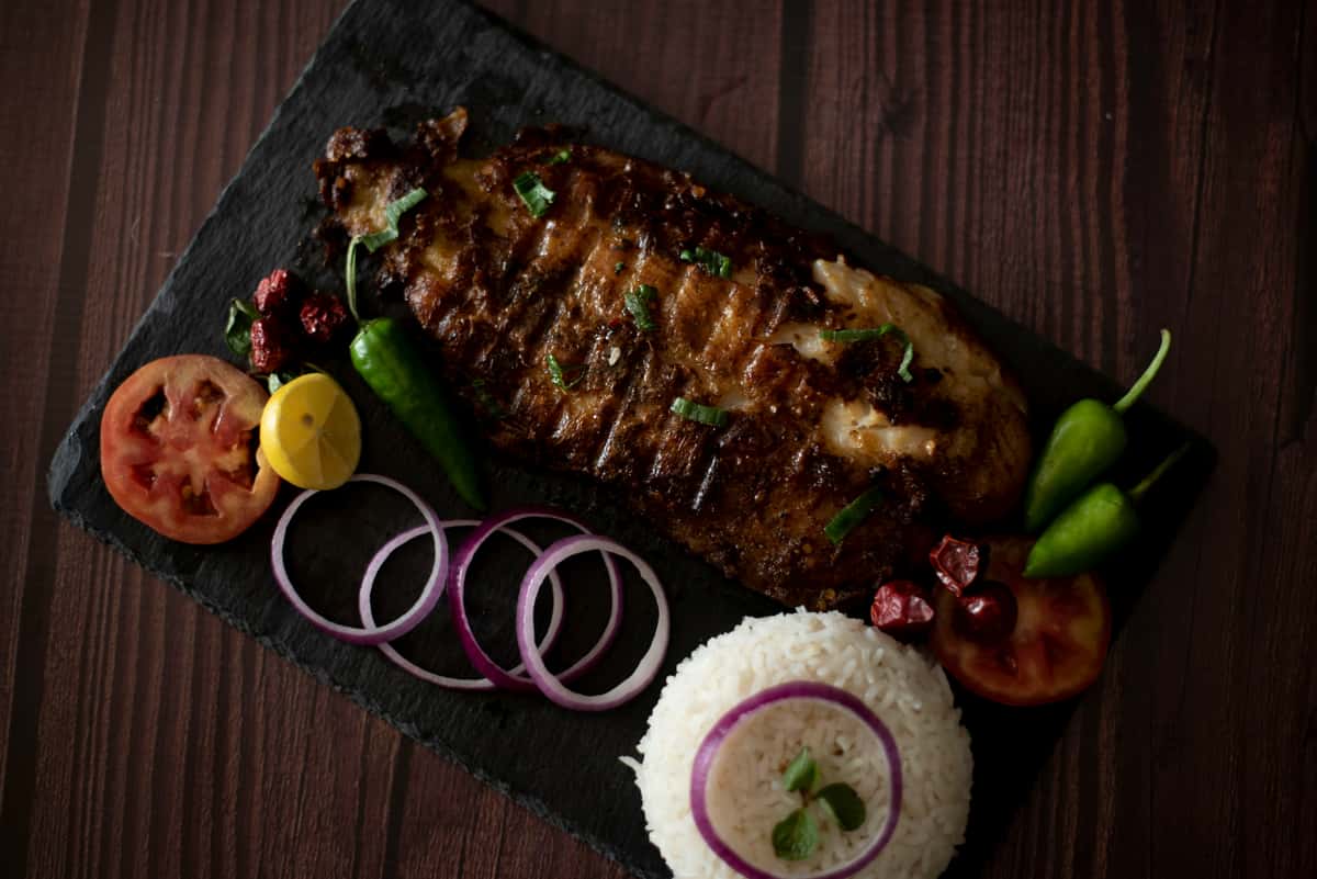 Grilled fish fillet served with white rice, sliced red onions, tomato, lemon, green chilies, and garnished herbs on a slate platter.