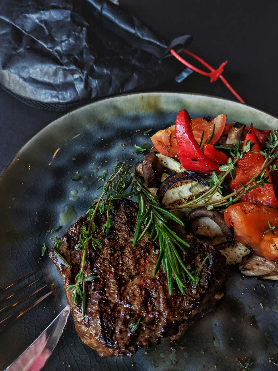 Grilled steak topped with rosemary, served with roasted vegetables including bell peppers, zucchini, and carrots on a dark plate.