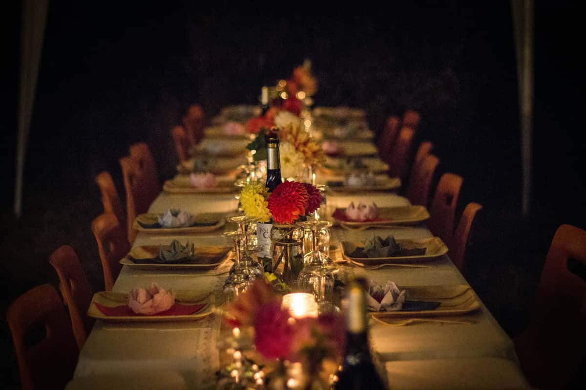 Romantic outdoor dining table set with wine, candles, folded napkins, and floral centerpieces under warm evening lighting.