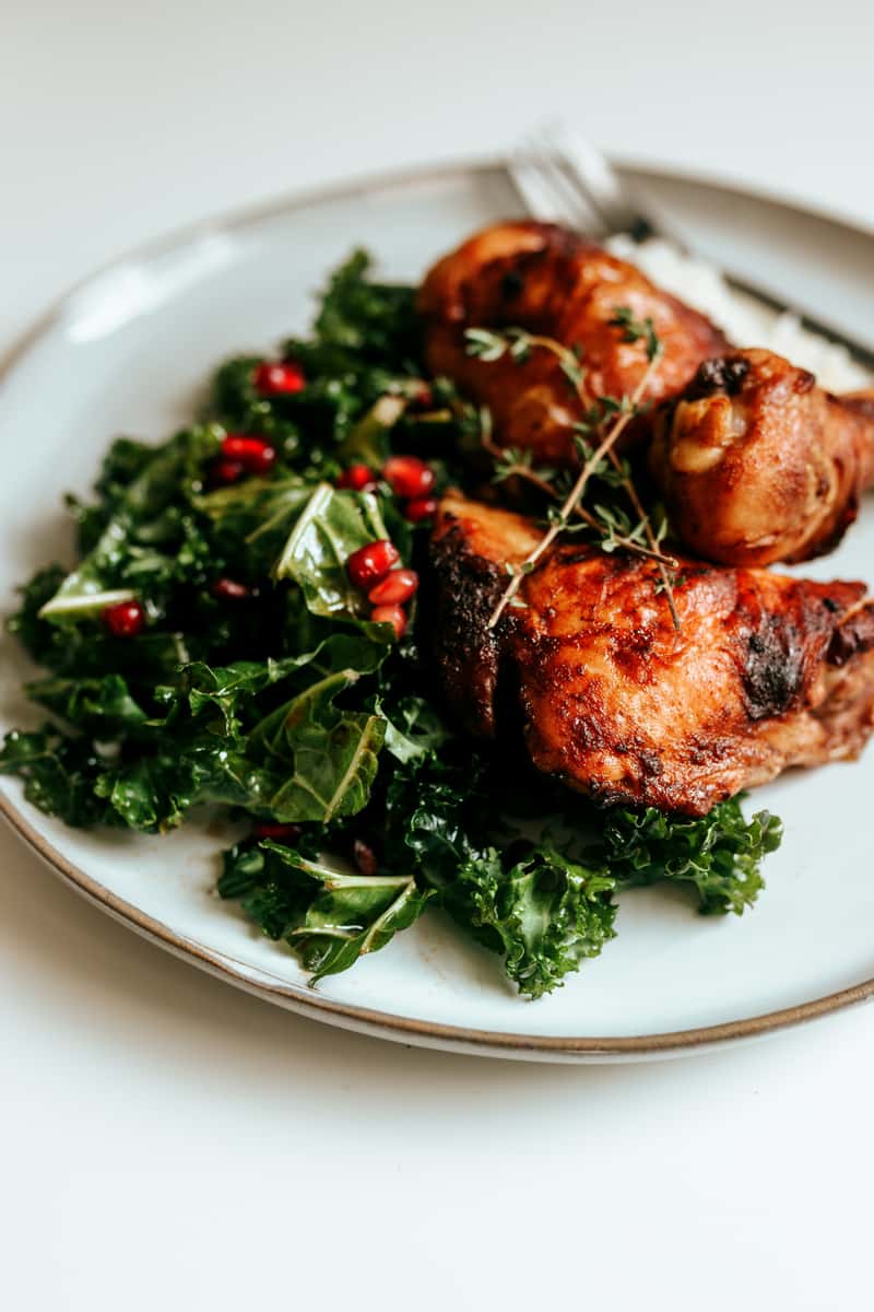 Roasted chicken with crispy skin served with kale salad topped with pomegranate seeds and fresh herbs on a white plate.
